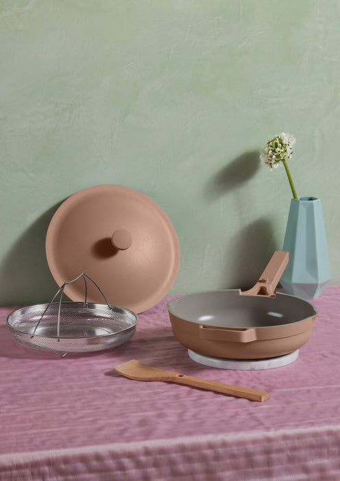 A pink Always Pan with spatula and steamer basket on a pink table