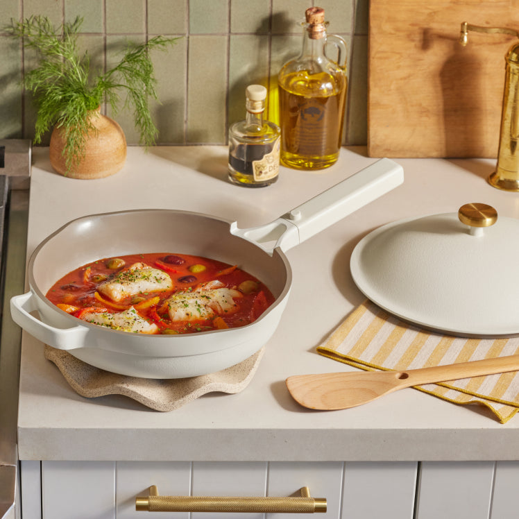 A white Always Pan with a tomato-based fish dish on a counter with its lid, spatula, and olive oil nearby